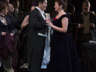 WNO La traviata Kang Wang Alfredo Linda Richardson Violetta and WNO Chorus Photo credit Betina Skovbro 2934 Betina Skovbro Alfredo and Violetta are looking intensely at each other, smiling and touching hands.