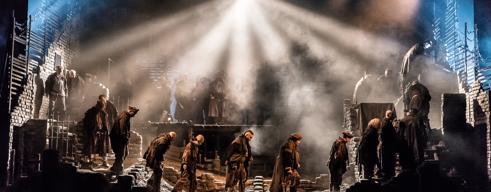 A dozen men wearing dark clothes walk somberly in line, across the dimly lit stage