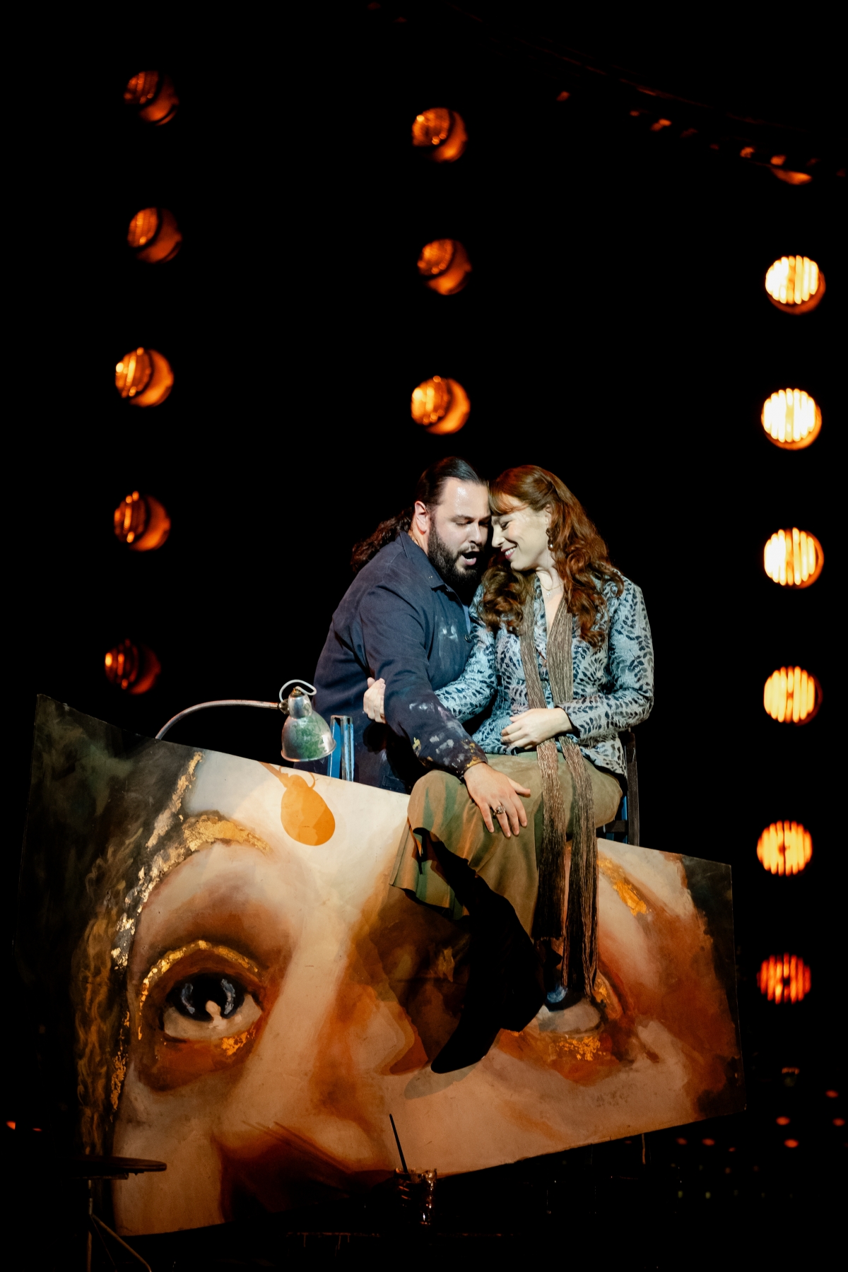 WNO Tosca Andrés Presno Cavaradossi Natalya Romaniw Tosca Photo Credit Dafydd Owen 400 Cavaradossi and Tosca share a moment of closeness and affection. Cavaradossi stands behind his painted fresco singing. Tosca sits on top of the fresco, her legs dangling over it. She smiles at Cavaradossi, clutching his arm while he holds her knee.