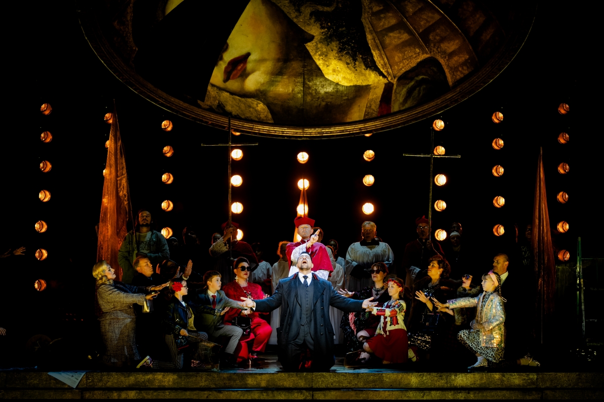 WNO Tosca Cast of Tosca Photo Credit Dafydd Owen 1004 Scarpia is on his knees at the centre of the stage. He wears a suit and formal jacket and raises his face and palms to the sky. Behind him is a priest, and a crowd of fashionable people and choral singers surround him, their arms reaching towards him. Many spotlights glow with light, and the golden dome hangs above.