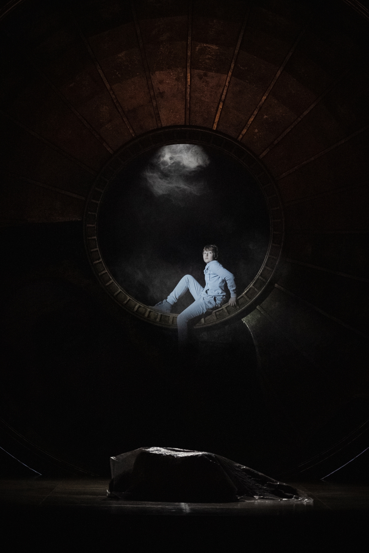WNO Tosca Cast of Tosca Photo Credit Dafydd Owen 2063 A young boy in a hoodie and jogging bottoms sits at the centre of the golden dome in semi-darkness, a spotlight shining on him. On the ground is a body covered by a large plastic sheet.