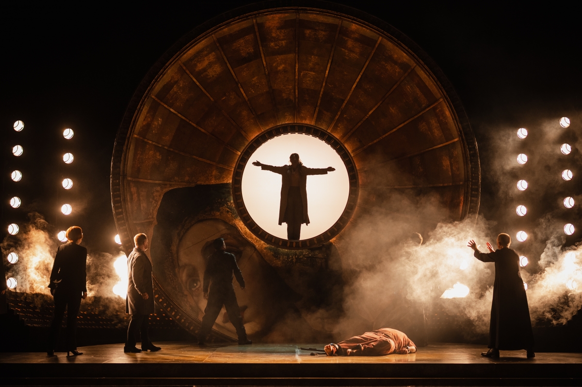 WNO Tosca Cast of Tosca Photo Credit Dafydd Owen 880 Tosca stands at the centre of the golden dome with her arms outstretched. Only her silhouette is visible against bright lighting. Below, Cavaradossi lies dead on the ground and various people look up at her.