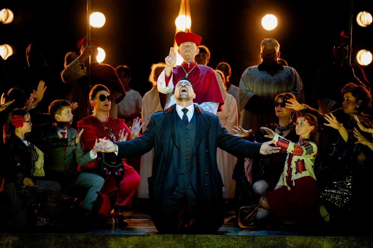 WNO Tosca Dario Solari Scarpia Cast of Tosca Photo Credit Dafydd Owen 1008 Scarpia is on his knees at the centre of the stage. He wears a suit and formal jacket and raises his face and palms to the sky. Behind him is a priest, and a crowd of fashionable people and choral singers surround him, their arms reaching towards him.