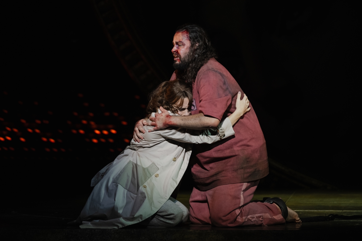 WNO Tosca Natalya Romaniw Tosca Andrés Presno Cavaradossi Photo Credit Dafydd Owen 2455 Tosca and Cavaradossi hold each other close. Cavaradossi wears a prisoner's outfit and is covered in blood and dirt. Tosca is on her knees and presses her head against Cavaradossi's chest.