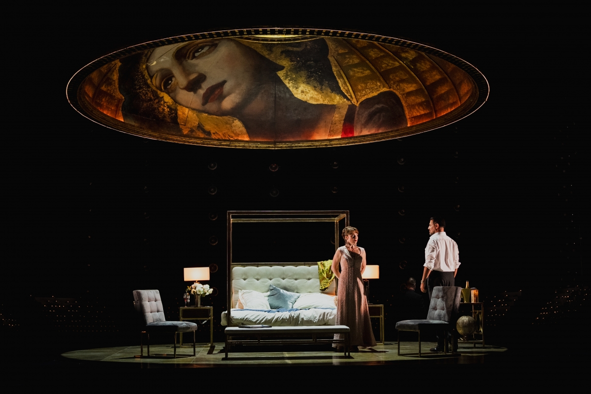 WNO Tosca Natalya Romaniw Tosca Dario Solari Scarpia Photo Credit Dafydd Owen 1201 Tosca and Scarpia are in Scarpia's room. Tosca, wearing her sparkling showcase gown, stands against the corner of his four-poster bed, an anxious expression on her face. Scarpia faces her. The golden dome with Mary Magdalene painted on to it hovers above them like a roof.