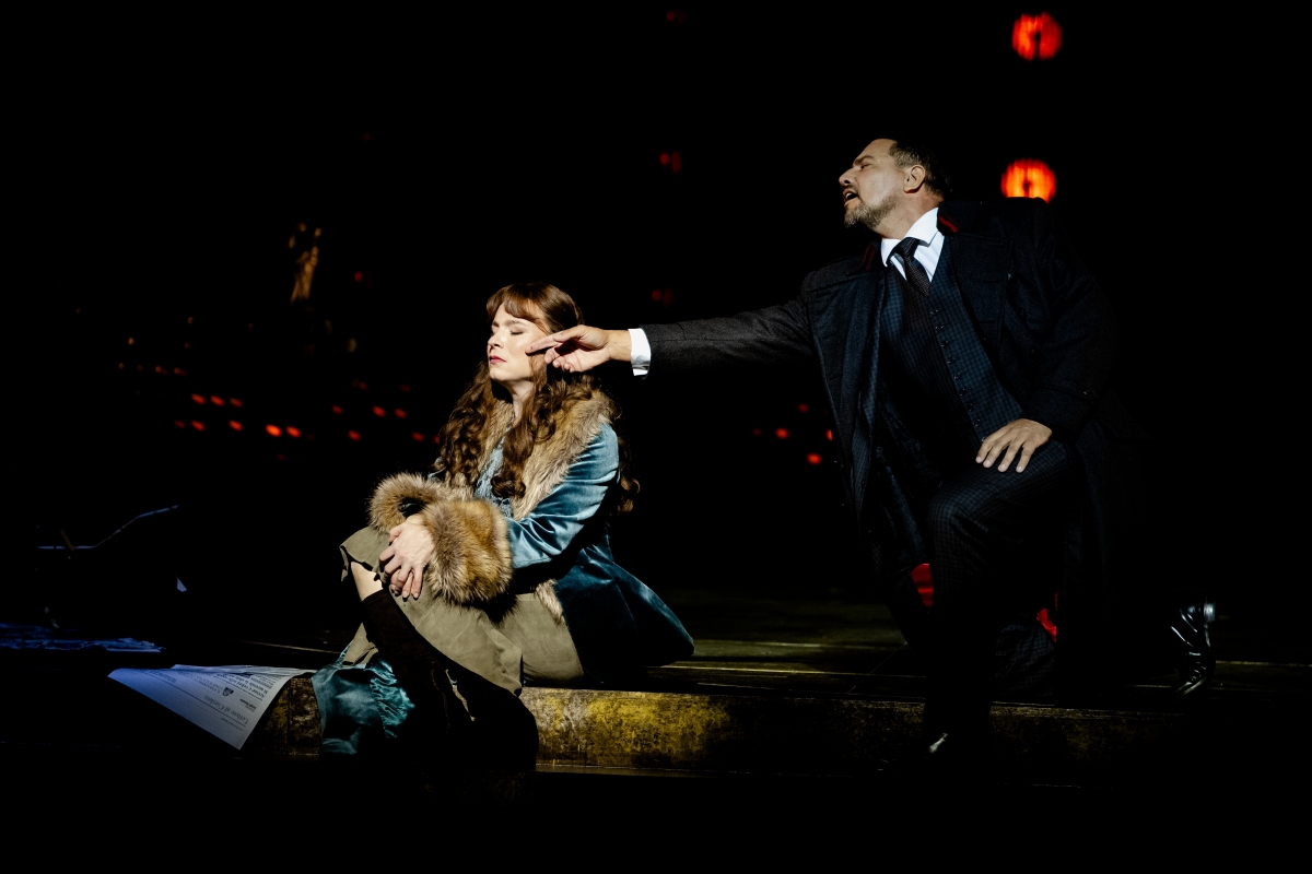 WNO Tosca Natalya Romaniw Tosca Dario Solari Scarpia Photo Credit Dafydd Owen 813 Tosca sits near the edge of the stage, wearing a velvet jacket with a fur collar and cuffs. Scarpia is down on one knee, reaching towards Tosca and brushing her cheek with his finger. She appears unhappy, facing away from him with her eyes closed.