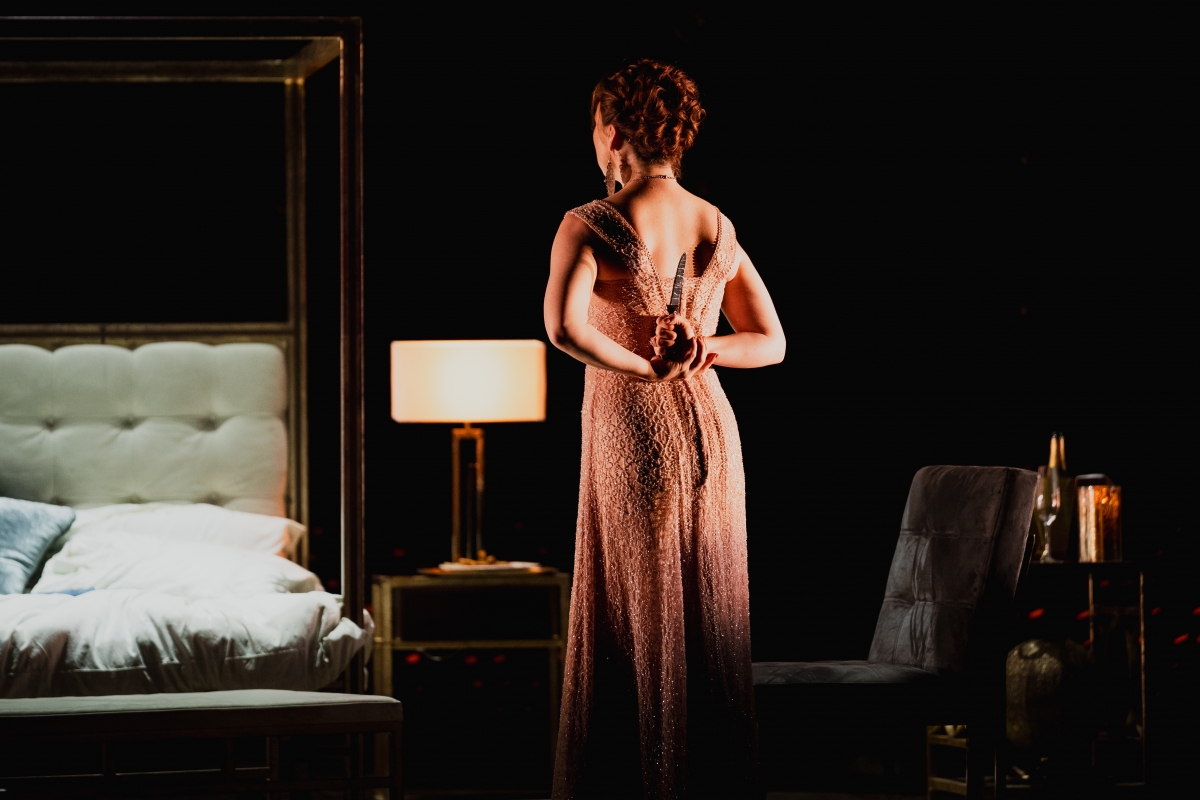 WNO Tosca Natalya Romaniw Tosca Photo Credit Dafydd Owen 1832 Tosca stands in Scarpia's bedroom facing away from the audience. She wears her sparkly showcase dress, and holds a steak knife behind her back.