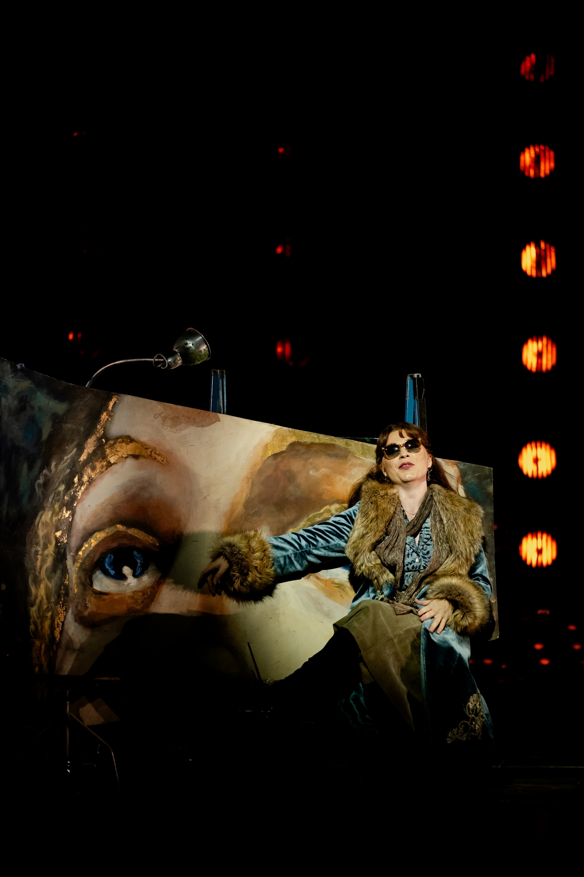 WNO Tosca Natalya Romaniw Tosca Photo Credit Dafydd Owen 514 Tosca sits in front of the fresco that Cavaradossi is painting, looking glamorous in a velvet jacket with a fur collar and cuffs. She also wears sunglasses, and gestures with her right hand to the fresco.
