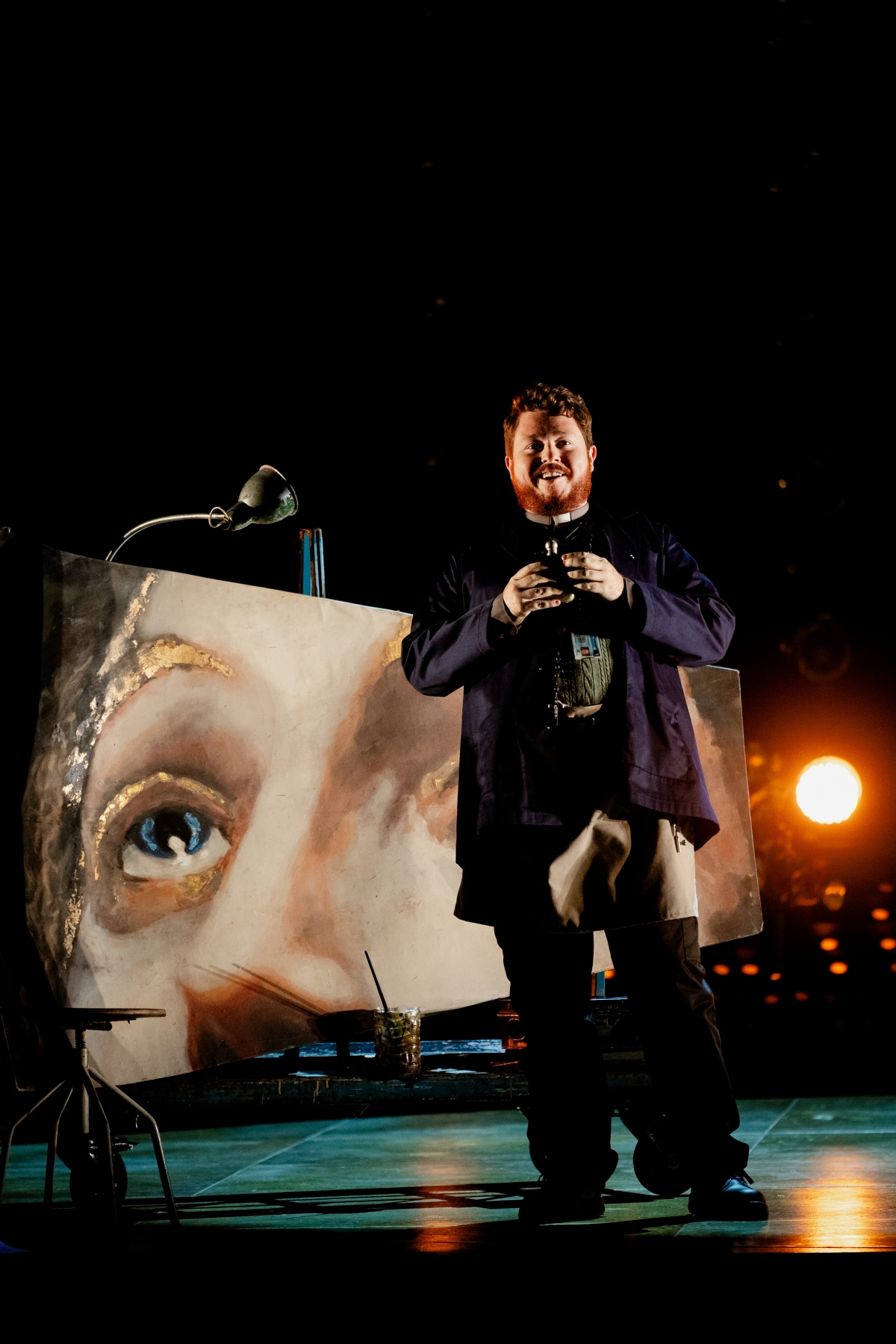 WNO Tosca Ross Fettes The Sacristan Photo Credit Dafydd Owen 589 The Sacristan stands facing the audience with a grin on his face, holding a vial of alcohol. Behind him is the fresco that Cavaradossi has been painting.