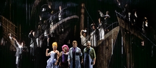 The Old Woman, Cunégonde, Candide and the Captain stand together centre stage, singing. The Old Woman holds up her arm in a gesture of determination. Behind, the WNO Chorus appear to stand upon animated boats, singing and waving handkerchiefs to the audience.