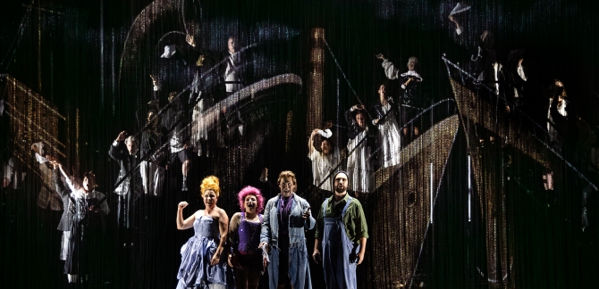 The Old Woman, Cunégonde, Candide and the Captain stand together centre stage, singing. The Old Woman holds up her arm in a gesture of determination. Behind, the WNO Chorus appear to stand upon animated boats, singing and waving handkerchiefs to the audience.
