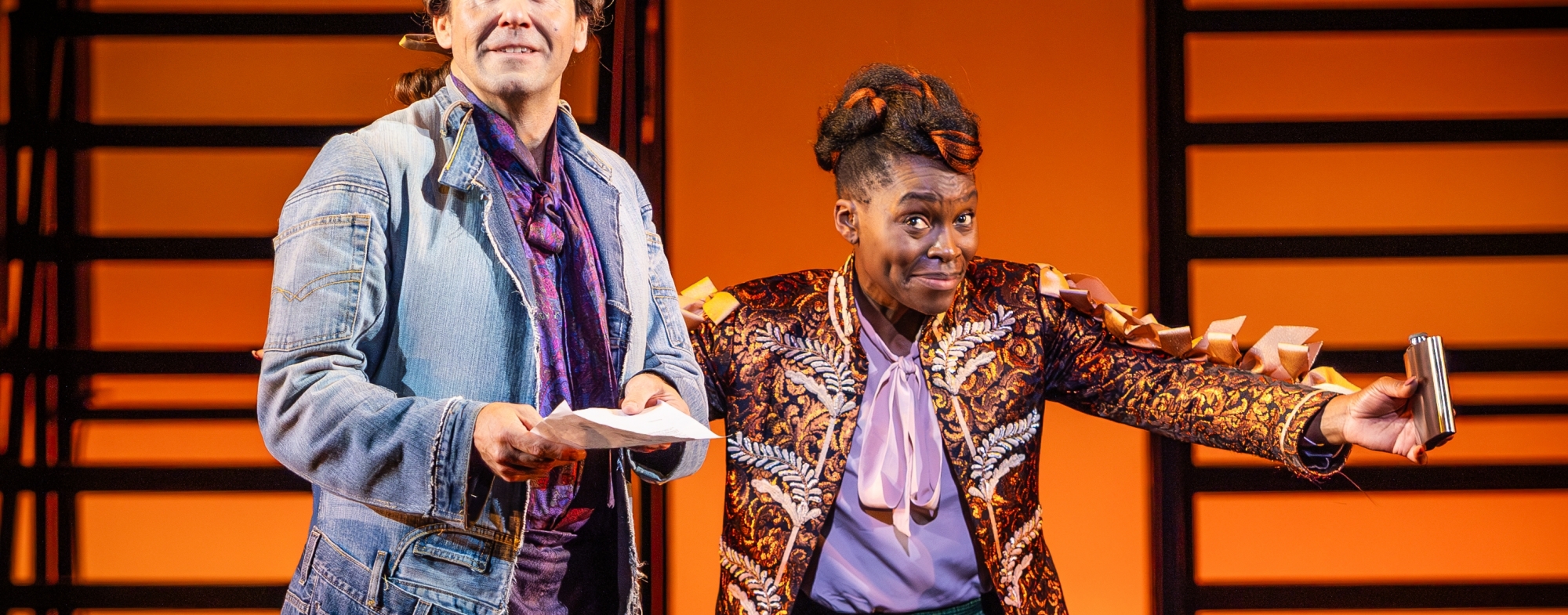 WNO Candide Ed Lyon Candide Rakie Ayola Pangloss Narrator Sage Beggar photo credit Craig Fuller 1173 Candide looks out to the audience with a hopeful smile. He wears a silk shirt and a long jacket made of jeans, and holds a piece of paper in his hands. Beside him is the Narrator/Pangloss, who has one hand on his back while the other clutches a hip flask.