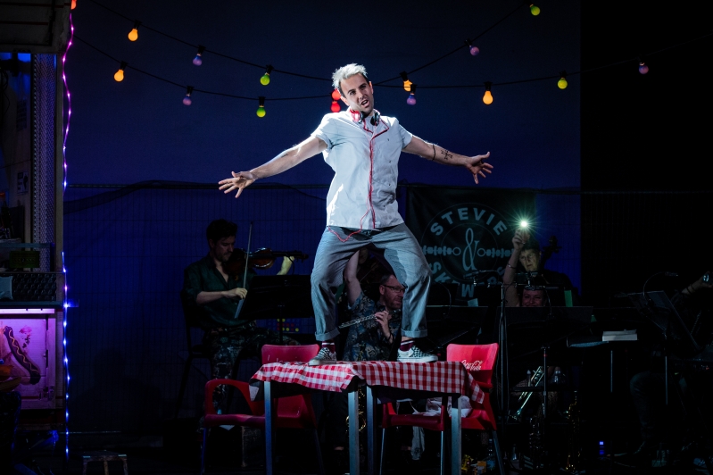 Man in chefs outfit standing on table singing with arms outstretched.