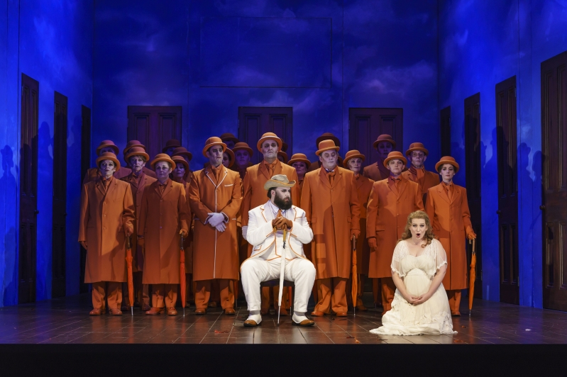 Woman in white kneeling at front, man sat in white, chorus standing in orange behind.