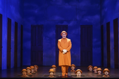 Man in orange clothes stands while other heads with orange bowler hats poke through the floorboards.