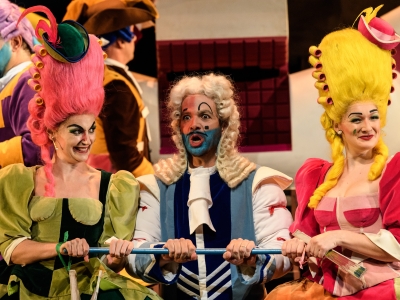 WNO La Cenerentola Aoife Miskelly Clorinda Giorgio Caoduro Dandini Heather Lowe Tisbe Photo credit © Jane Hobson 7623 Jane Hobson Clorinda and Tisbe, smiling, stand either side of Dandini, who appears surprised.
