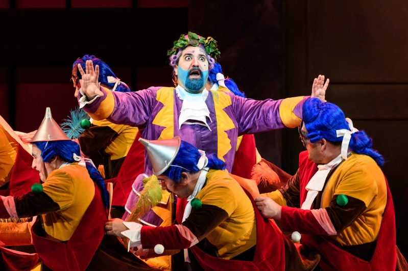 Don Magnifico appears shocked, his hands in the air. He is surrounded by the WNO Chorus, dressed in red, yellow and blue outfits.