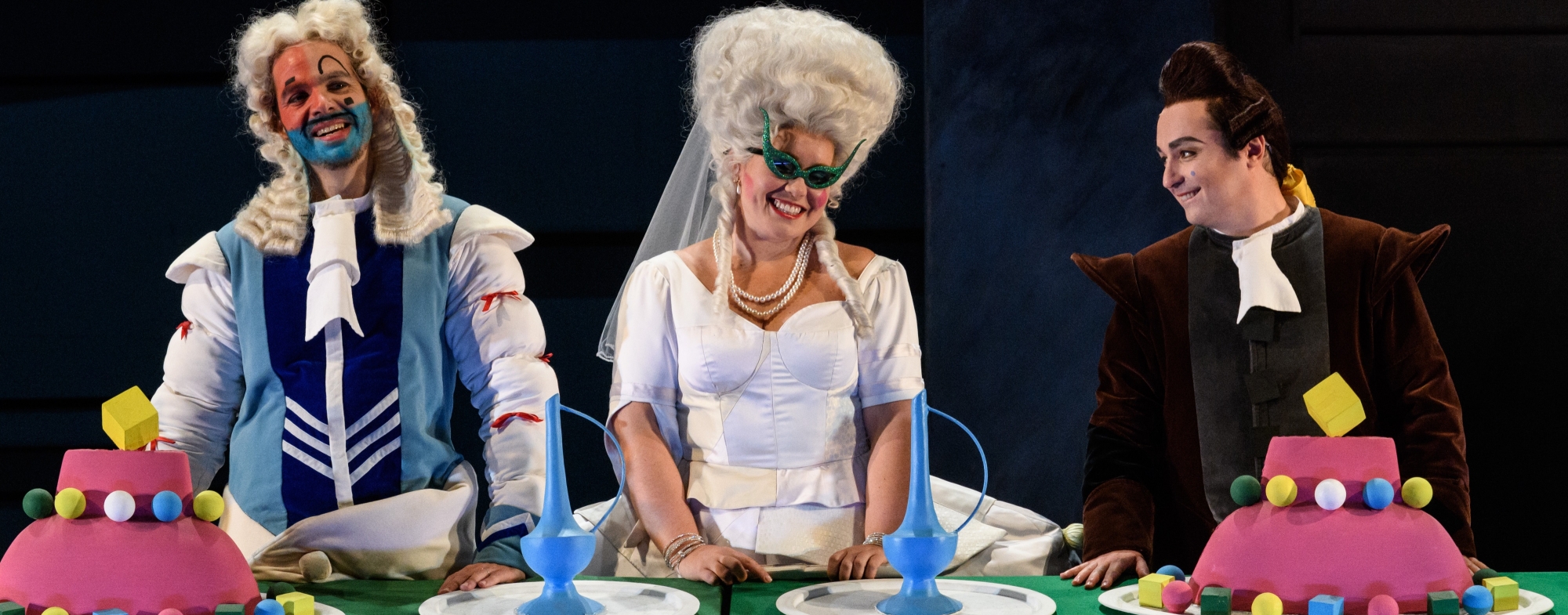 Dandini, Angelina and Don Ramiro sit happily at a table. There are brightly coloured cakes in front of them.