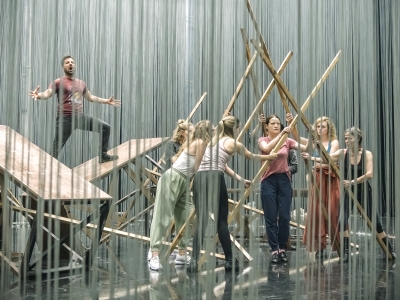 a group of performers arrange beams of wood into a triangular tunnel, as a man stands on two tables