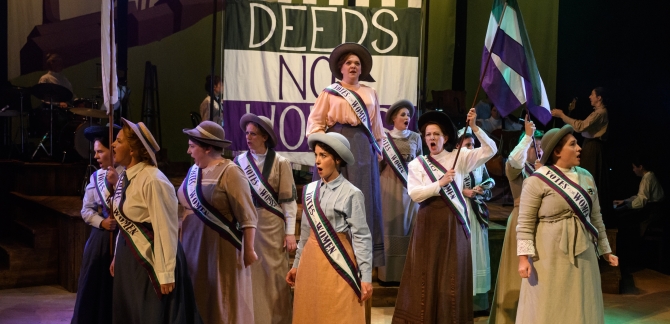 WNO Cast Of Rhondda Rips It Up! Photo Credit © Jane Hobson 2524 Image shows the women's choru sin traditional dress, marching in front of a green, white and purple Deeds Not Words banner. They are waving green, white and purple flags and wearing Vote for Women Sashes