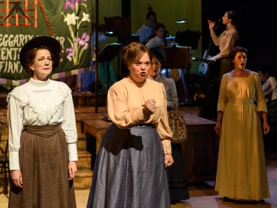 Sybil is dressed in a white frilled blouse and brown skirt, Lady Rhondda is dressed in a beige blouse and blue skirt. They look out to the distance. Lottie and Helen Archdale stand slightly behind in the darker light watching on. 