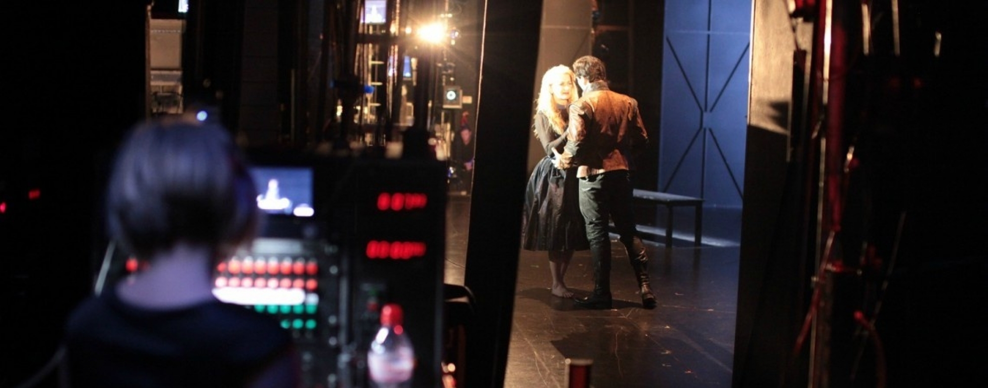 WNO Backstage Test Image A Photo Grapher 2016 Taken from the wings we see a technician int he forefront of the image, a male and female performers on stage gaze intently at each other. Bright stage lighting.
