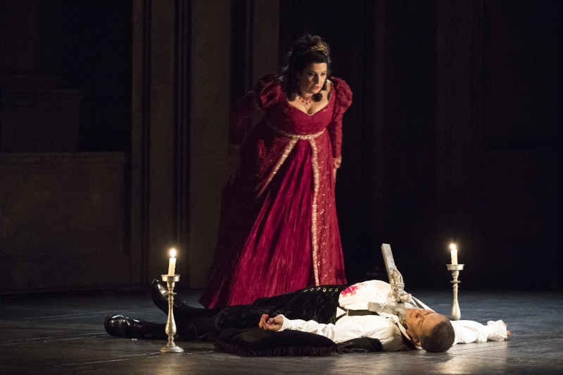 Man lies on floor between two candles with object protruding out of his neck, woman stands looking. 