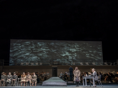 Wide shot of stage woman standing infront of desk whilst others are seated on the other side.