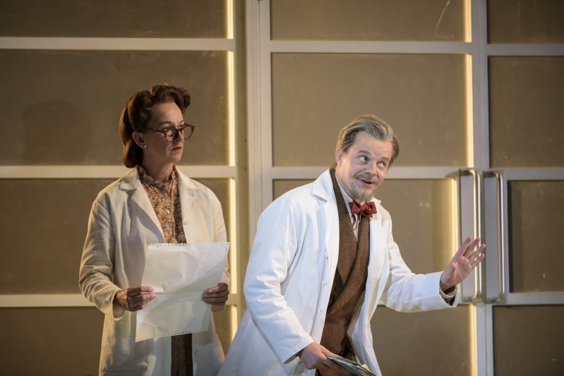 Man in white doctors coat gestrues raising his eyebrows whilst woman with paper stands behind him.