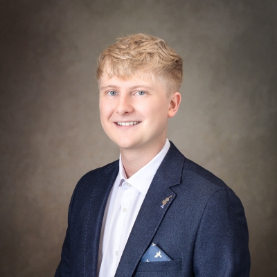 A headshot of Owain Rowlands, who is sat on a slight angle smiling at the camera. He wears a smart shirt and suit jacket. A tiny violin pin badge is attached to one of the jacket's lapels.