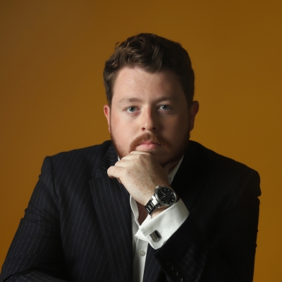 A headshot of Ross Fettes, who looks straight at the camera with a serious expression on his face. He wears a smart shirt with cufflinks and a pinstripe suit jacket, and sits on a backwards-facing chair. He leans on this chair with one elbow, his hand supporting his chin.