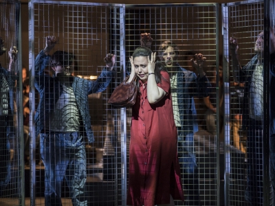 Woman holds her head between her hands as men lean against metal fence behind.