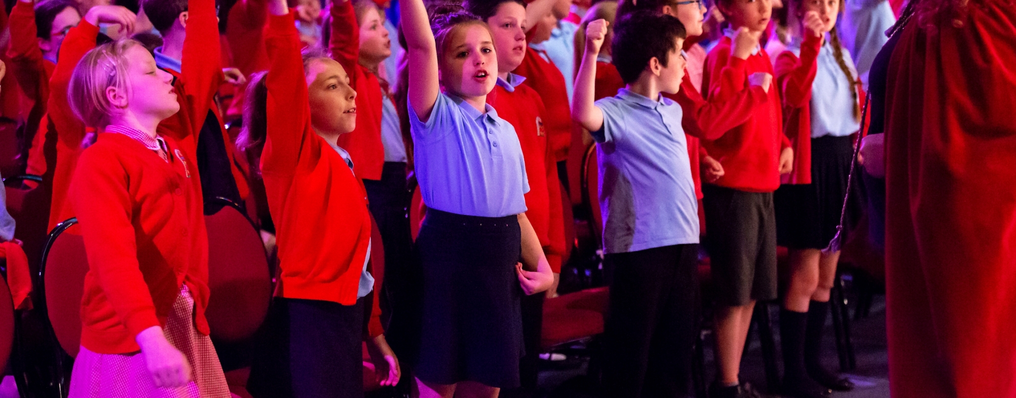 WNO Schools Concert Llandudno Venue Cymru May 21 2018 credit Paul Sampson 48 Paul Sampson 2018 A large group of Primary School children in red and blue uniforms sing, some with arms in the air. Black box rehearsal room setting.