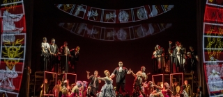 Massive roulette wheels frame the stage as a man in black stands on a table surrounded by party-goers in vibrant pink and red outfits