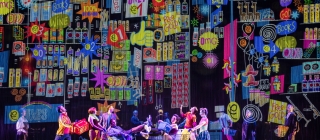 A group of colourfully dressed dancers parade full shopping trolleys before a wall of colourful bright signs