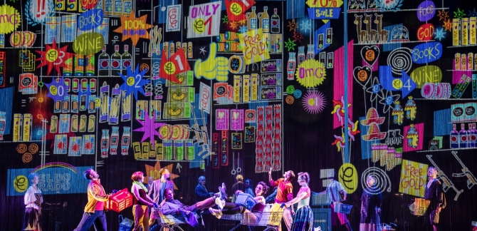 A group of colourfully dressed dancers parade full shopping trolleys before a wall of colourful bright signs
