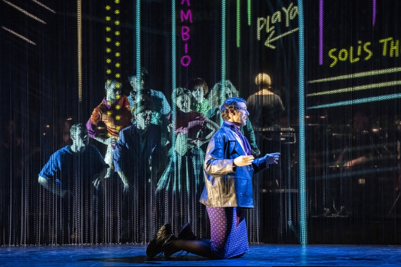 WNO Candide Mark Nathan Maximilian photo credit Johan Persson 07742 a man in a leather jacket and purple skirt kneels, as in the background a group of dancers watch on, engrossed