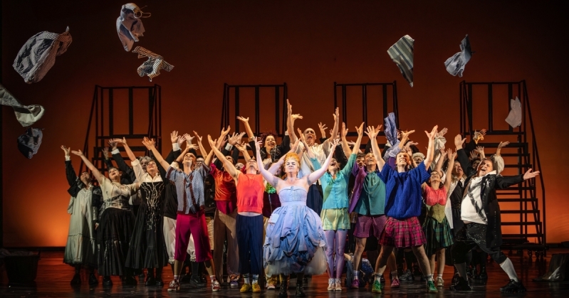 Candide Costumes Image 1 The Old Lady stands centre stage in her dress made of shirts, her arms in the air triumphantly. Behind her, the dancers and the WNO Chorus all do the same gesture, smiling gleefully. They have thrown multiple items of clothing into the air, which fly above the crowd.