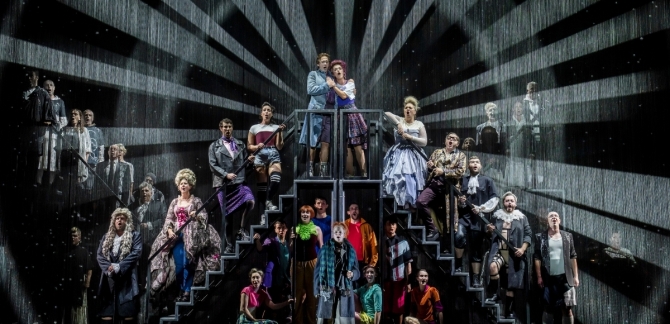The full cast of Candide stand on the steps of multiple adjoining staircases. Animated beams of light surround them as they sing.