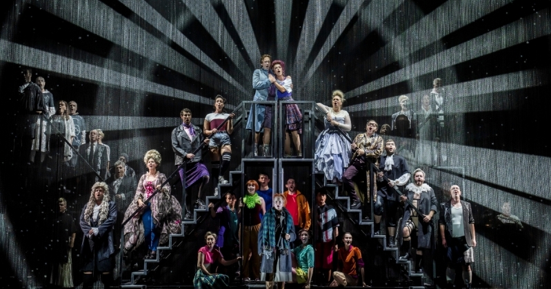 Candide Full Cast Photo The full cast of Candide stand on the steps of multiple adjoining staircases. Animated beams of light surround them as they sing.