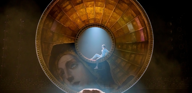 A huge golden dome featuring a fresco of Mary Magdalene hangs vertically above the stage. In semi-darkness, a bright light shines through a large hole in the dome's centre, where a young girl sits. The light illuminates a body in a bag on the floor downstage centre.
