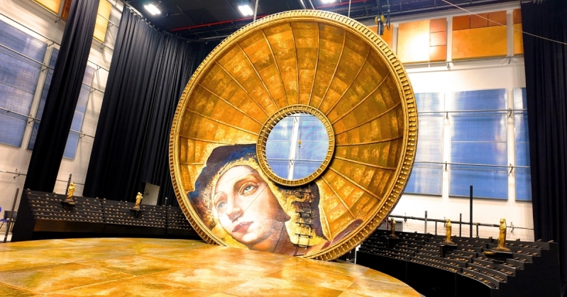 A wide backstage photo of the Tosca set. At the centre is a huge golden dome featuring a fresco of Mary Magdalene. Surrounding this is a semi-circular block covered in tiny lights which look like candles, along with small gold statues of religious figures.